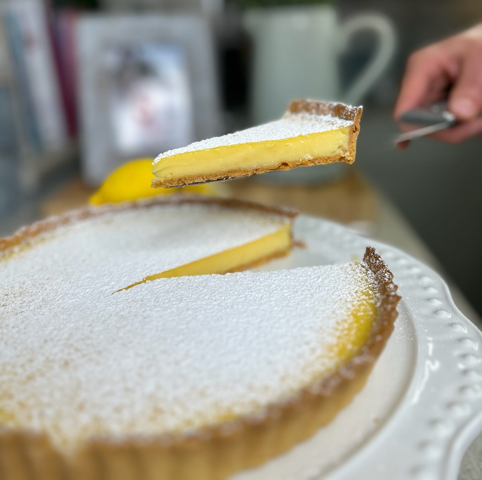 A lemon tart dusted with icing sugar on a cake stand with a slice cut on a knife lifted ready to be served