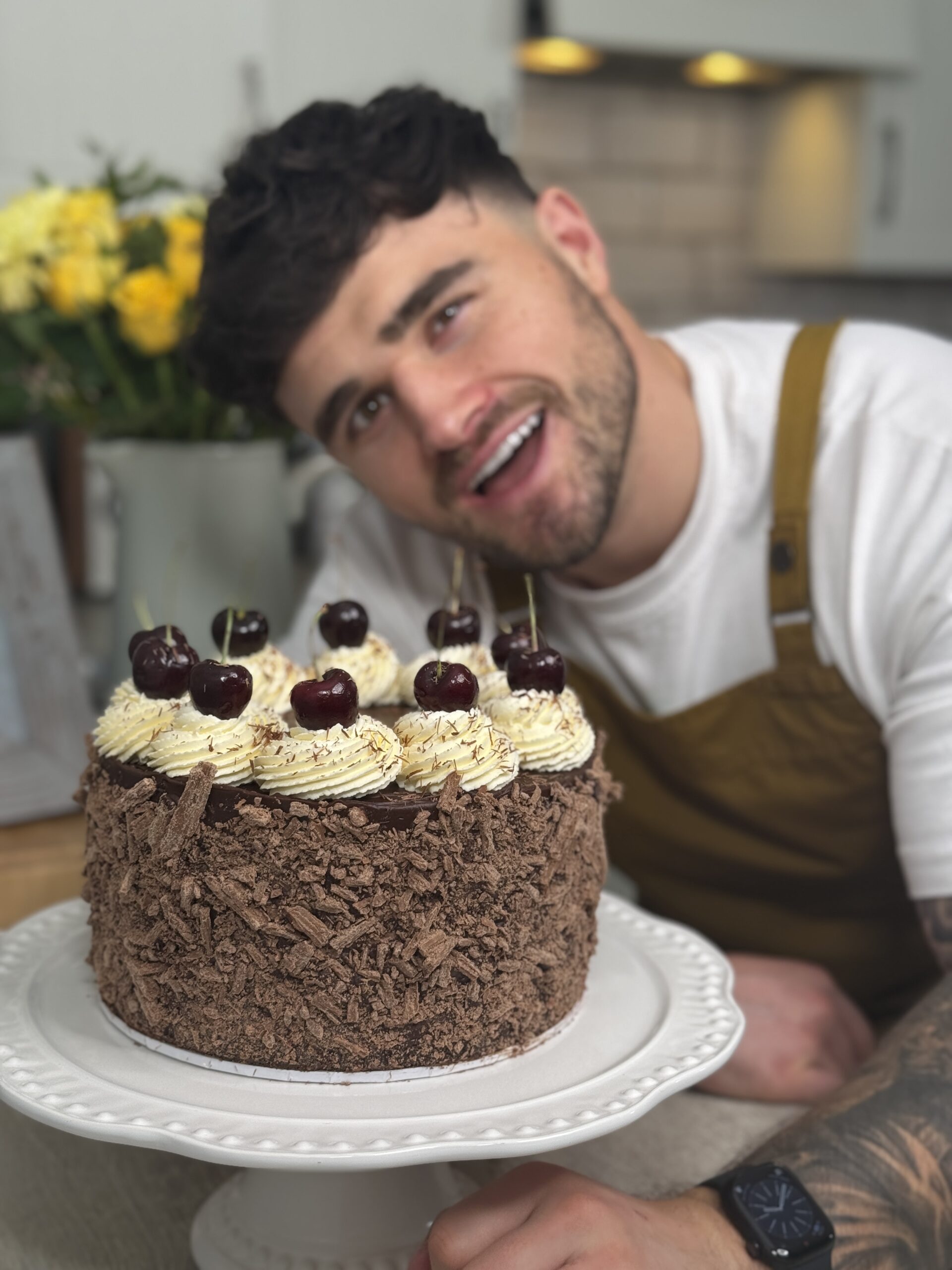 Matty smiling behind the finished cake that is coated in flakes of chocolate and topped with swirled cream and cherries